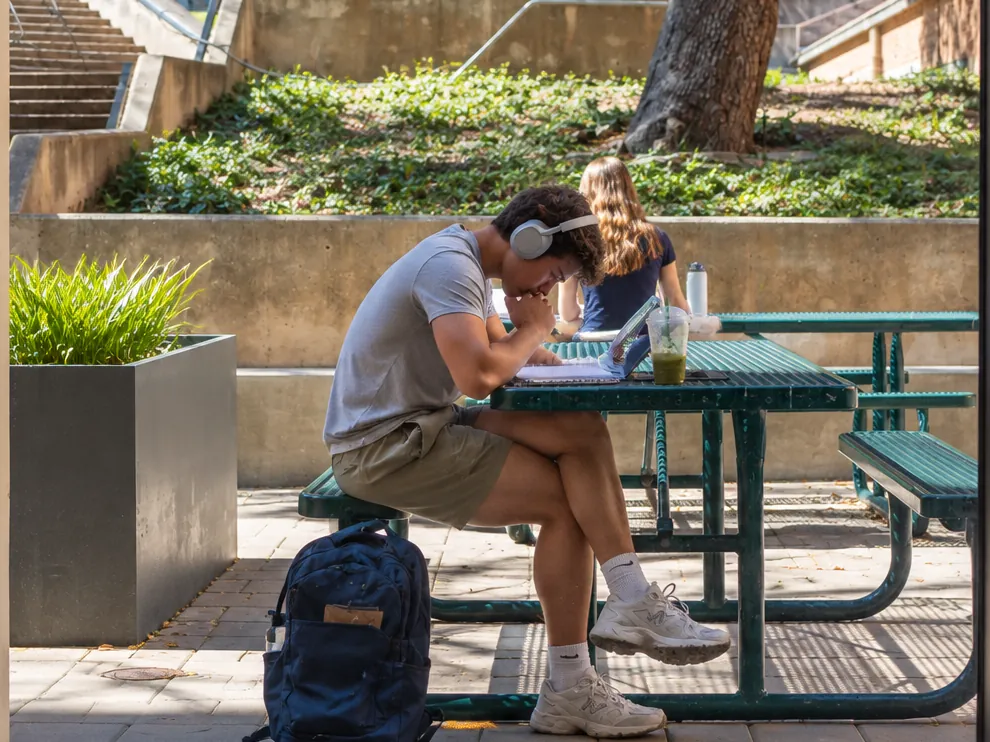 A student working through coursework at an outdoor campus table, with a notebook, laptop and backpack beside him
